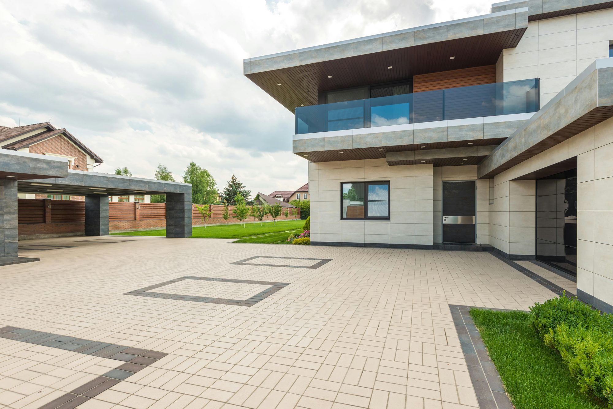 Modern two-story home with pale stone tile cladding, a glass-railed upper balcony, and deep cantilevered roof overhangs, set beside a wide paved motor court with a flat-roofed carport, a strip of lawn, and neighboring houses behind a brick and wood fence under a cloudy sky.