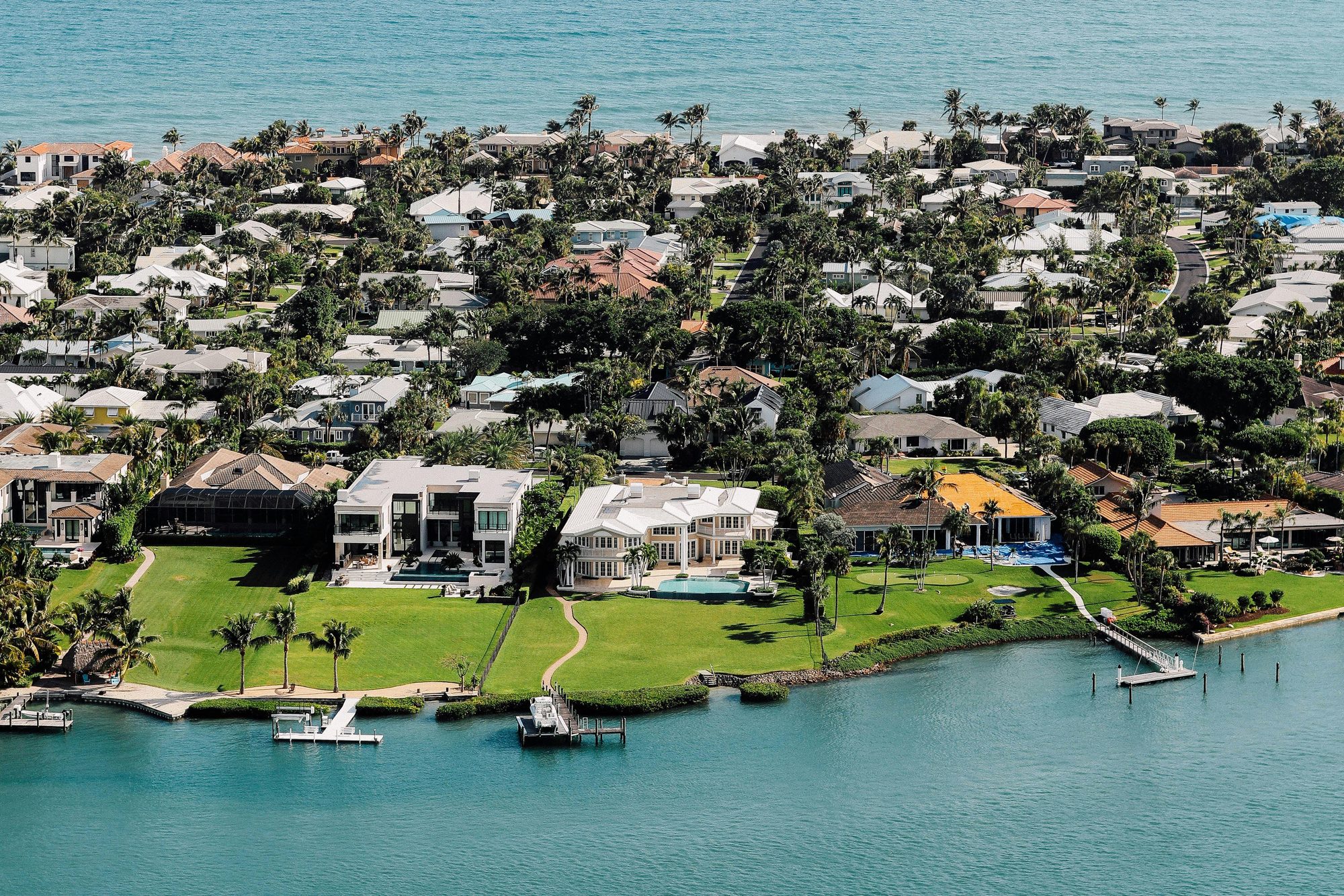 Aerial view of a waterfront neighborhood with a large white modern home and a white two-story home with a pool on wide green lawns along a turquoise bay, private docks and boats at the seawall, palm-lined streets of homes behind, and open water in the distance.