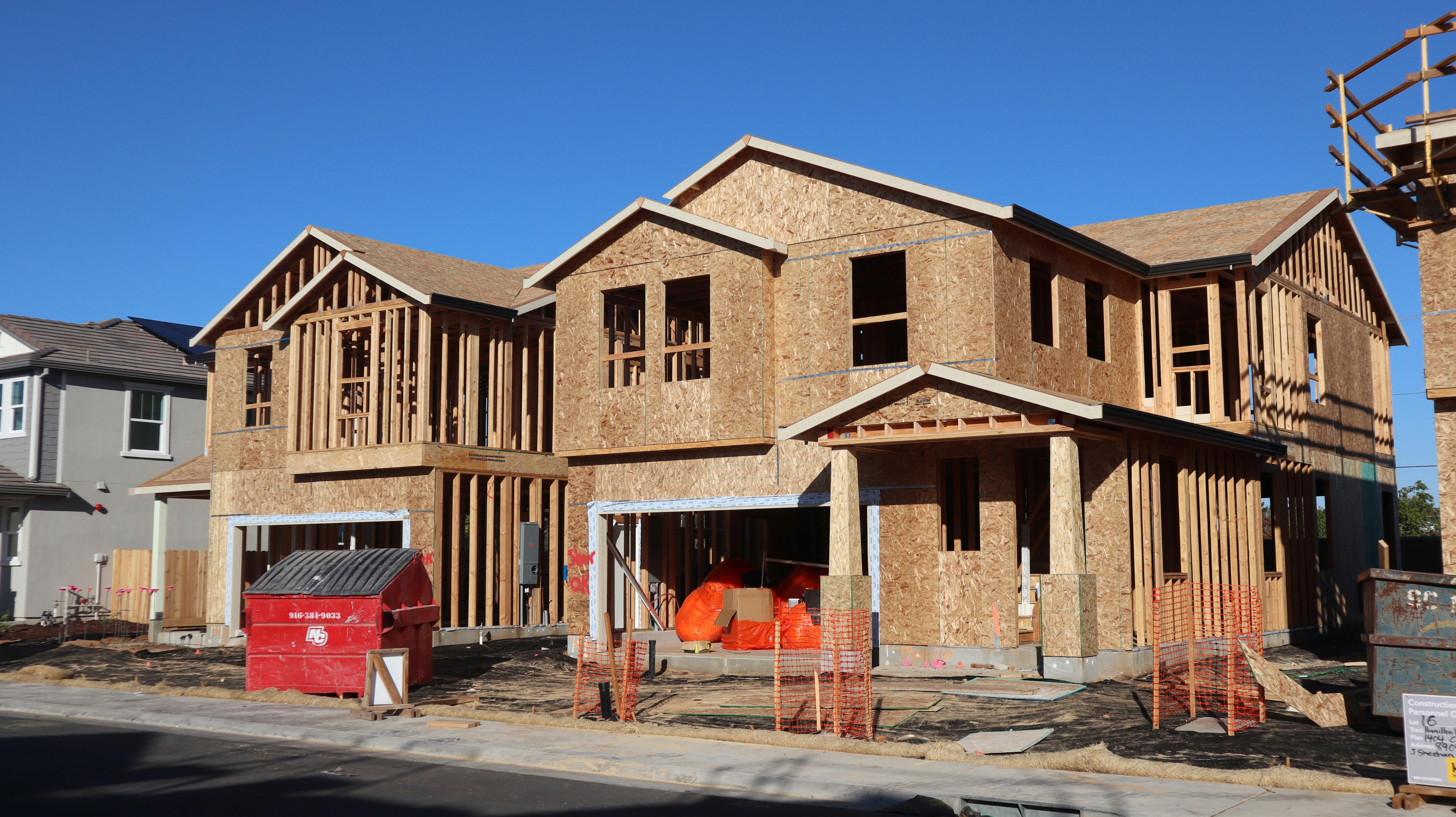 A large two-story house under construction with wood framing and sheathing, a red dumpster at the curb, and orange safety fencing along a new sidewalk under a clear blue sky.