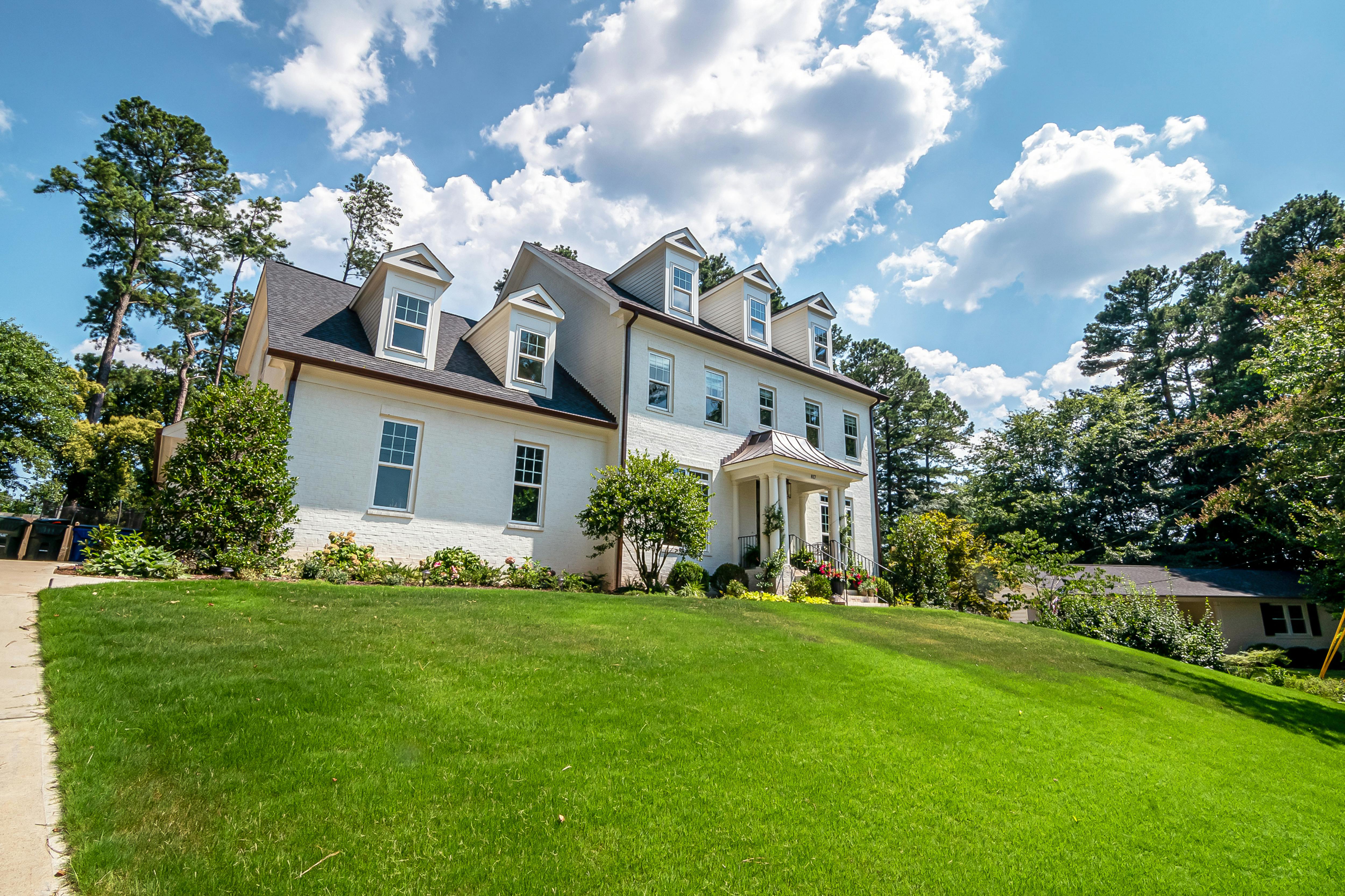 A white brick two-story house with dormers and a covered front entry on a sloped green lawn, with tall pines and a smaller older house beside it.