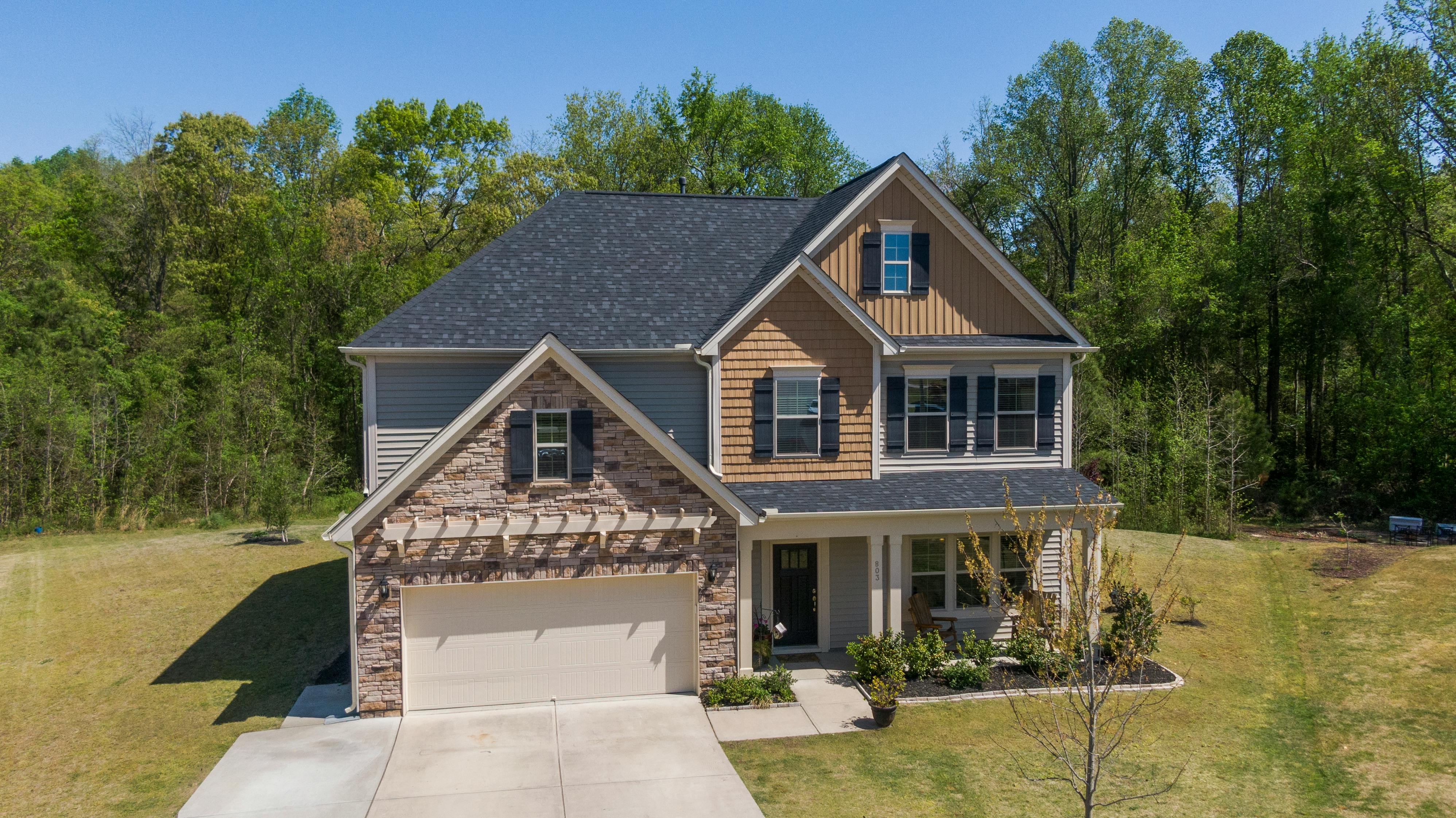 A two-story house with stone and shingle siding, dark shutters, a two-car garage, and a wide green lawn backed by tall trees.