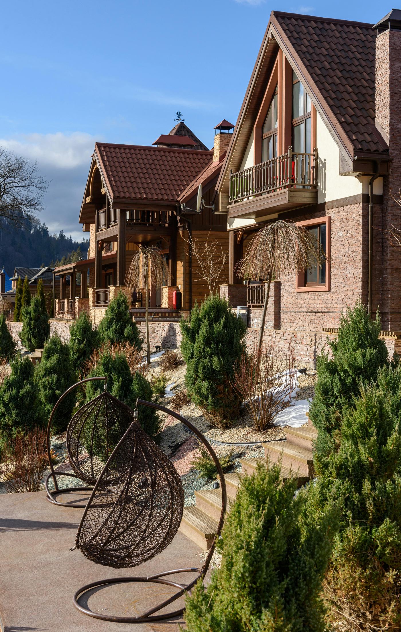 Two-story homes with brick and wood siding, steep tiled roofs, and upper balconies above a stepped stone retaining wall, wooden stairs, evergreen shrubs, and a hanging wicker chair under a blue sky.