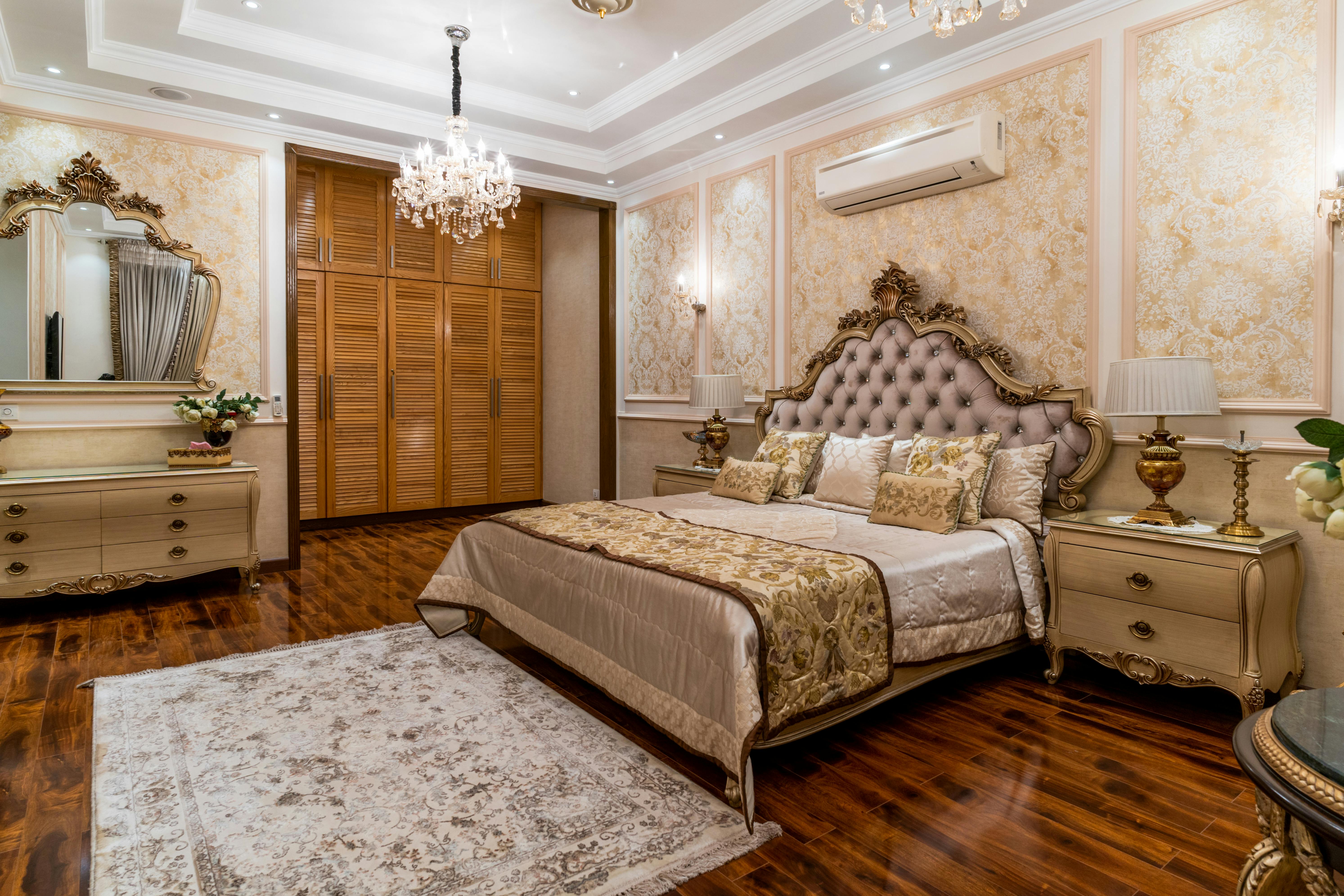 An ornate bedroom with a tufted headboard, gold-trimmed nightstands with lamps, a crystal chandelier, and dark hardwood floors under a patterned rug.