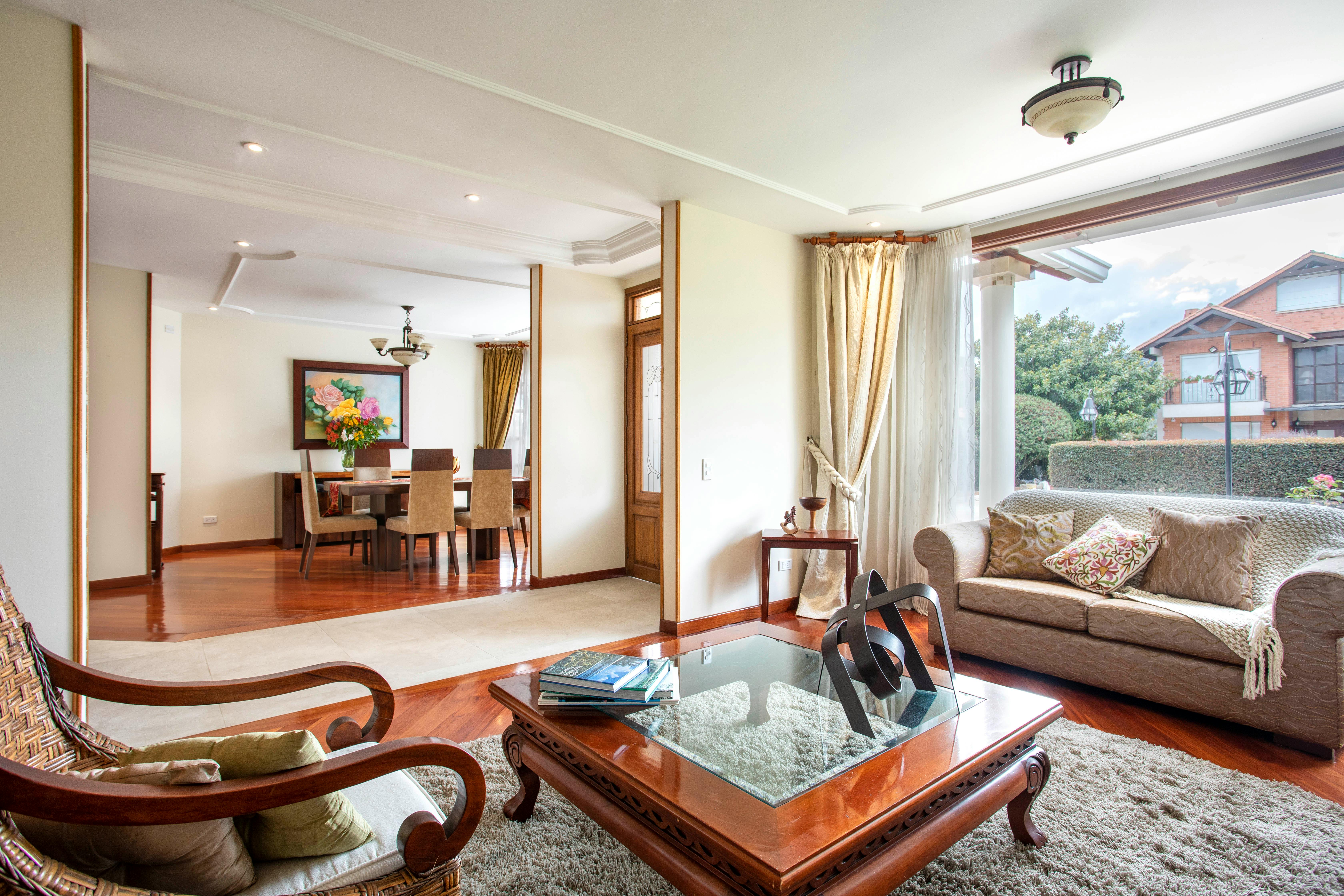 A traditional living room with a carved wood coffee table, a beige sofa beside a large window, and a dining room with a floral painting beyond.