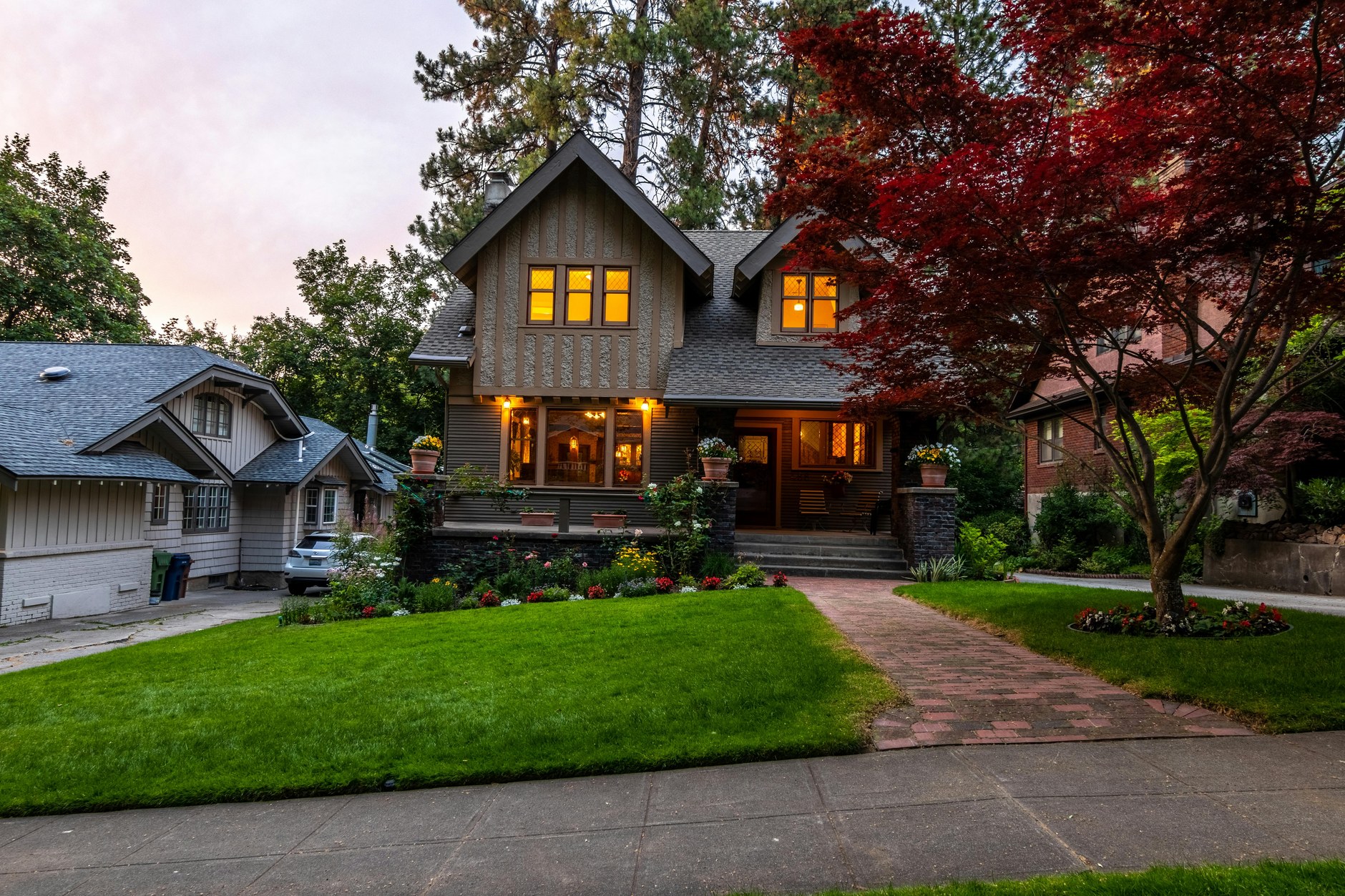 A two-story Craftsman cottage with stone porch, warm window light, and a red maple on a manicured front lawn at dusk.